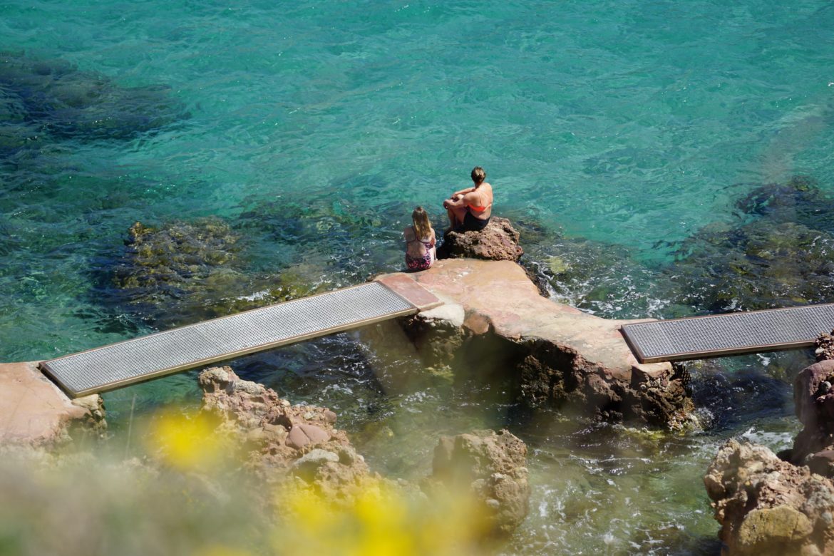 Two swimmers sit at the waters edge dipping their toes